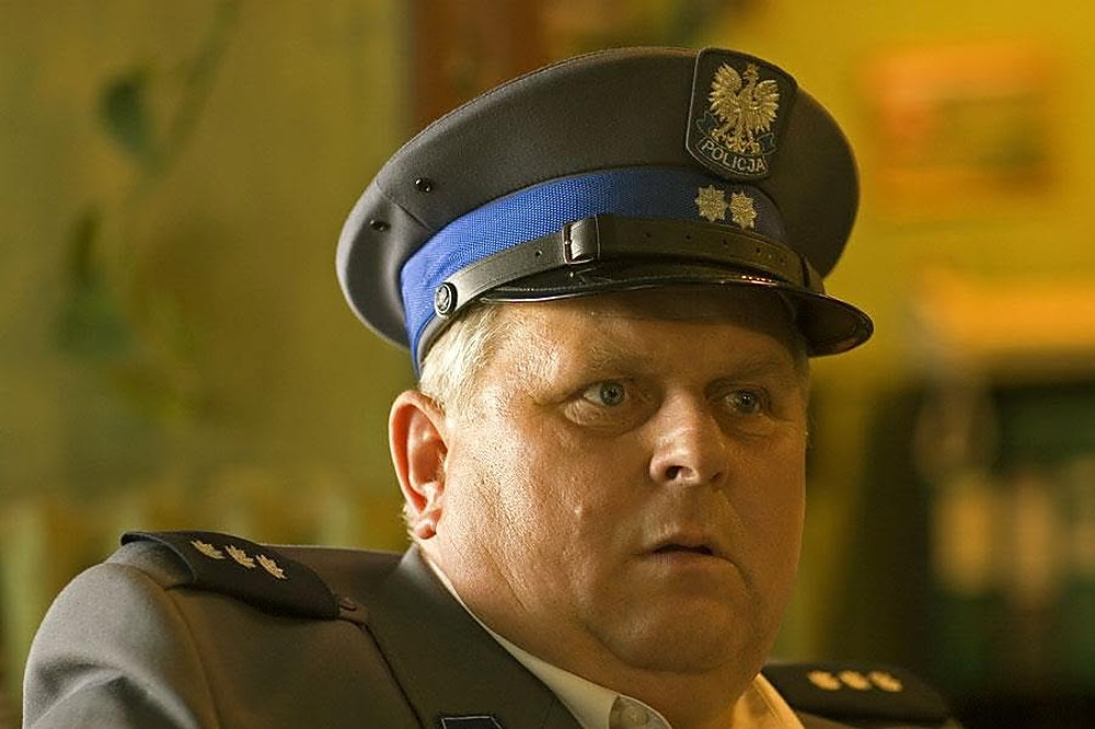 Aktor w policyjnej czapce z orzełkiem i mundurze z trzema gwiazdkami patrzy w bok z uniesionymi brwiami