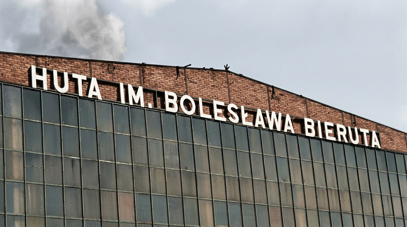 Ceglana ściana szczytowa hali hutniczej z wielkimi białymi literami HUTA IM. BOLESŁAWA BIERUTA i dymem nad dachem