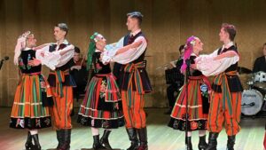 Pary w pasiastych strojach ludowych tańczą na scenie, w tle zespół muzyczny gali Być Polakiem