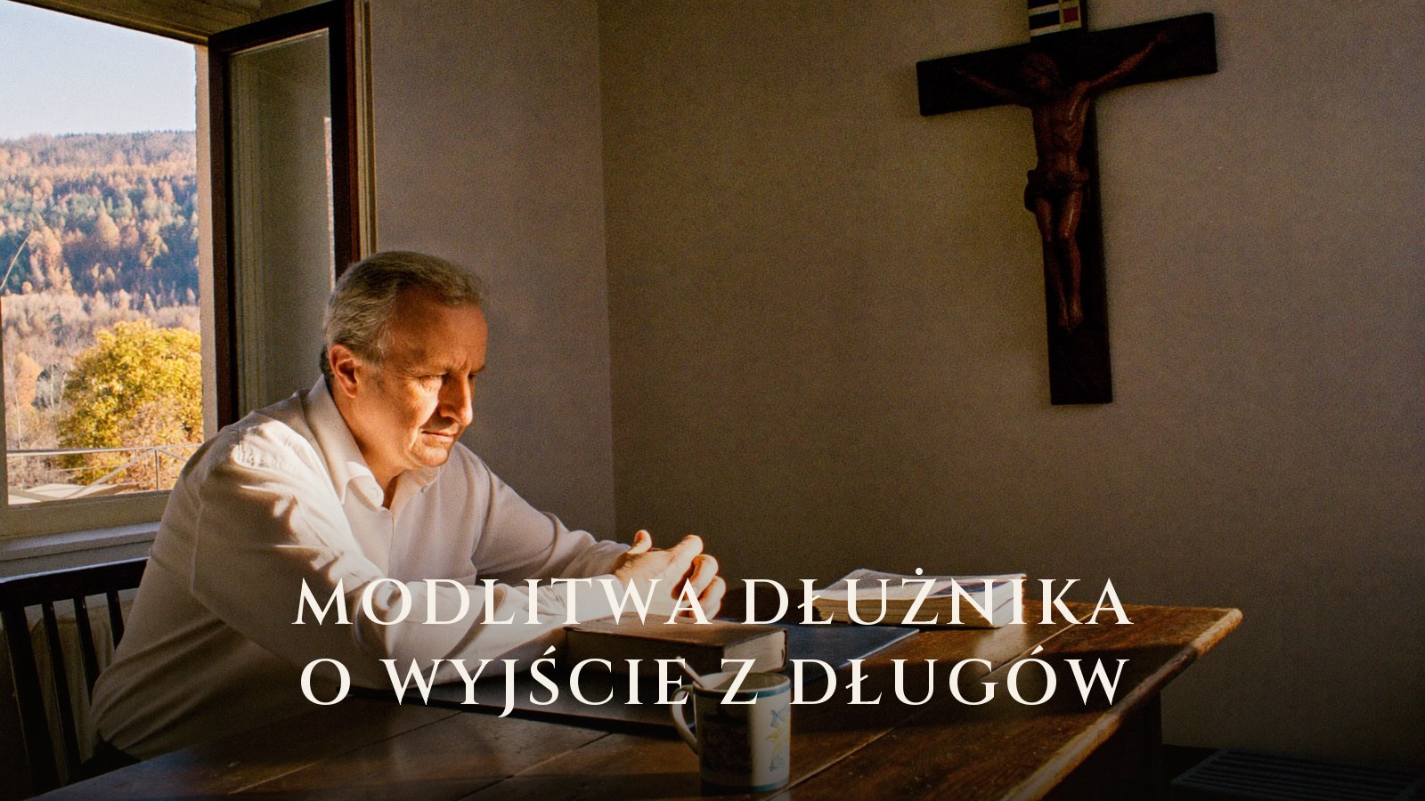 Mężczyzna modli się przy stole nad modlitewnikiem i rachunkami, na ścianie krucyfiks