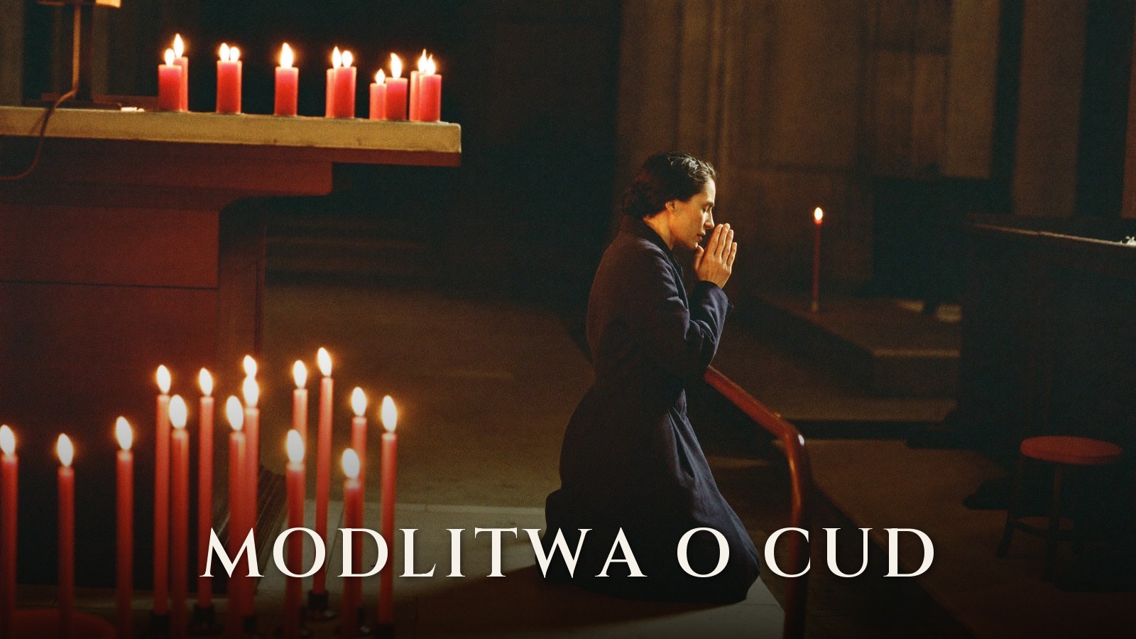Kobieta w granatowym płaszczu klęczy w kościele przed rzędem zapalonych czerwonych świec