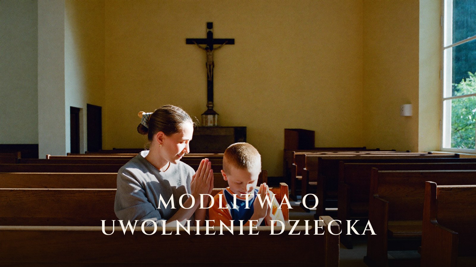 Matka z synem modlą się ze złożonymi dłońmi w ławce kościelnej, w tle krucyfiks nad ołtarzem