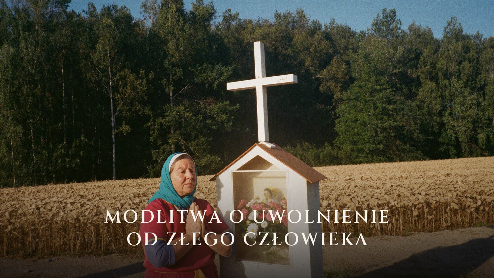 Starsza kobieta modli się przy przydrożnej kapliczce z krzyżem, w tle pole zboża