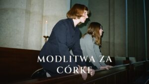 Matka i dorosła córka klęczą razem w ławce kościelnej, obok pali się świeca