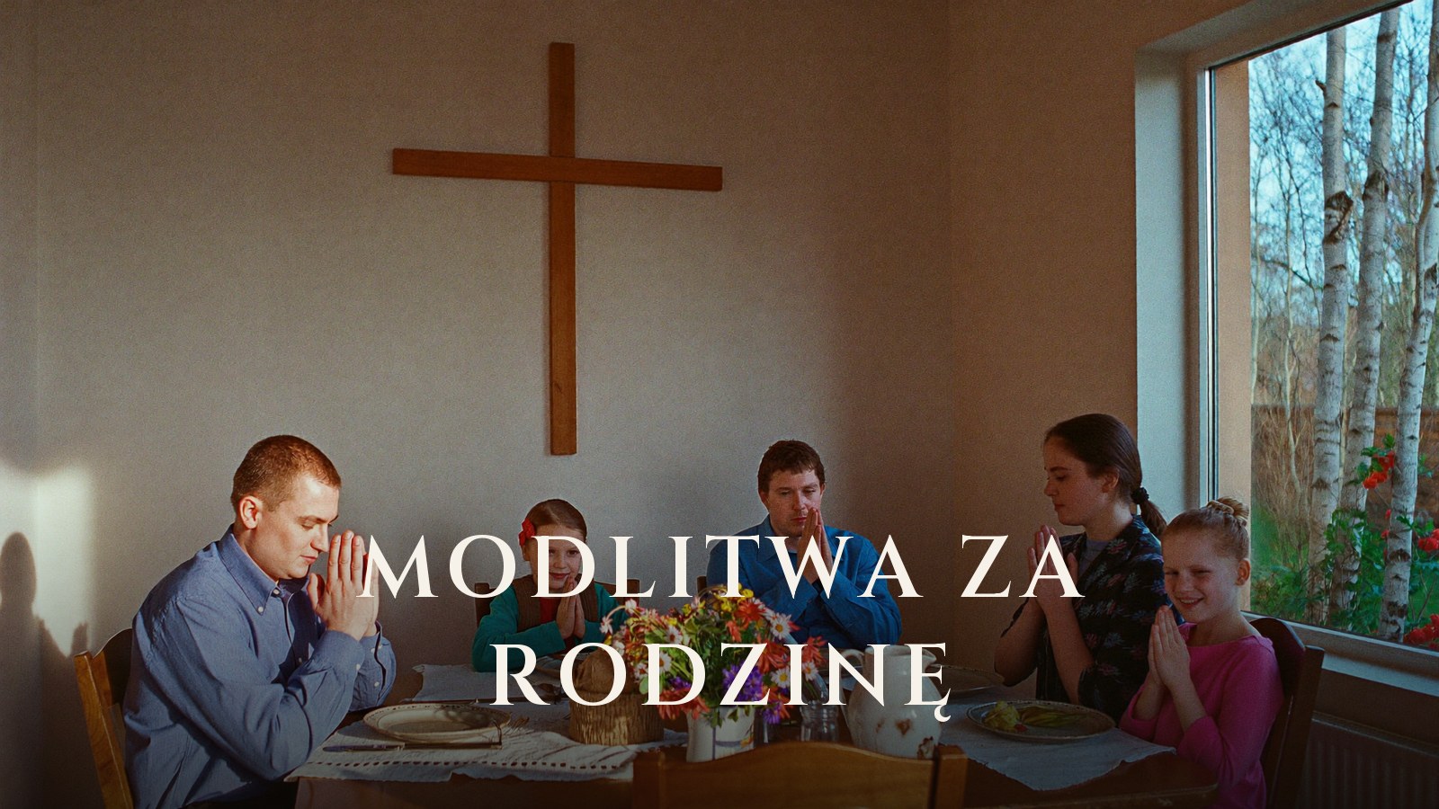 Rodzice z dwojgiem dzieci modlą się ze złożonymi dłońmi przy stole w domu, na ścianie drewniany krzyż