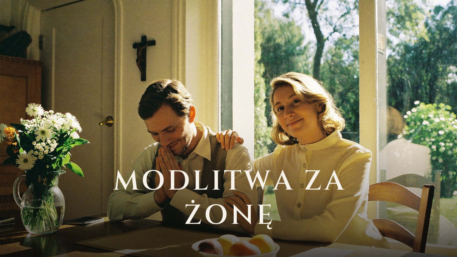 Mąż ze złożonymi dłońmi modli się przy stole, obok niego uśmiechnięta żona