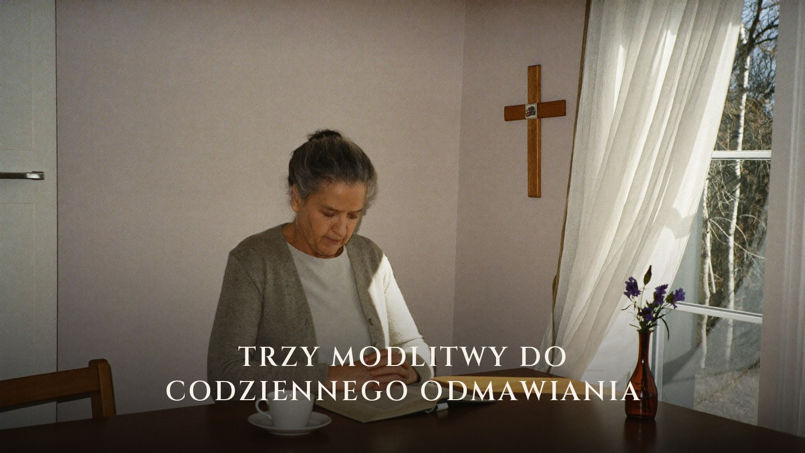 Starsza kobieta modli się nad otwartym modlitewnikiem przy kuchennym stole o poranku, na ścianie krzyż