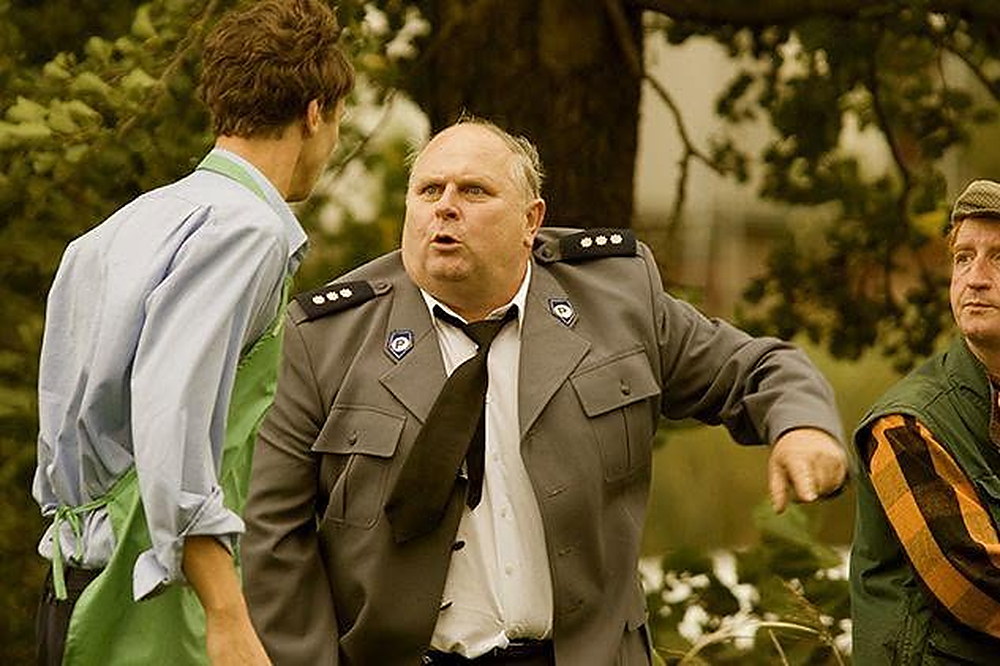 Komendant policji w mundurze tłumaczy coś mężczyźnie w zielonym fartuchu, obok stoi trzeci mężczyzna