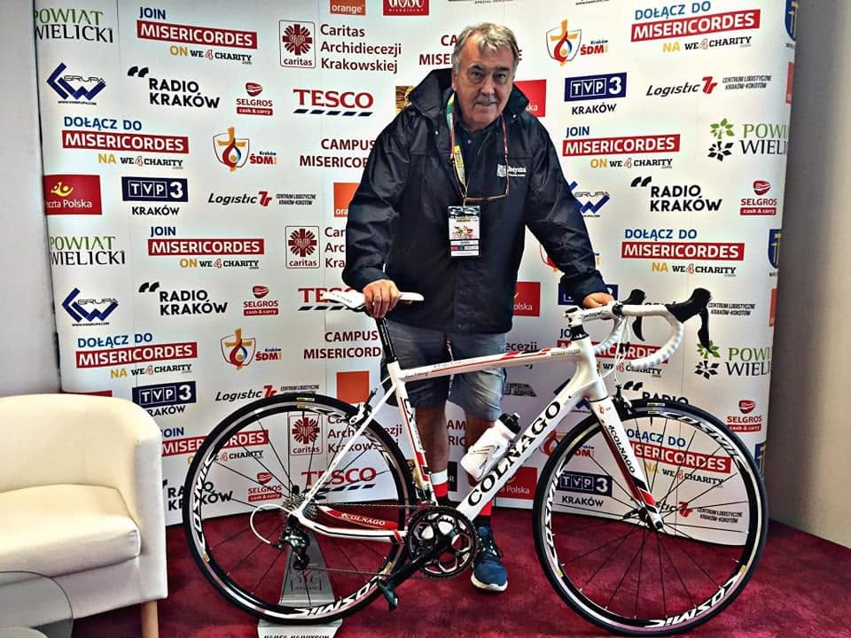 Włodzimierz Rezner w granatowej kurtce trzyma biały rower szosowy Colnago przed ścianką sponsorską akcji charytatywnej