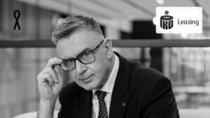 Andrzej Krzemiński w ciemnym garniturze poprawia okulary, siedząc w przeszklonym wnętrzu biurowym