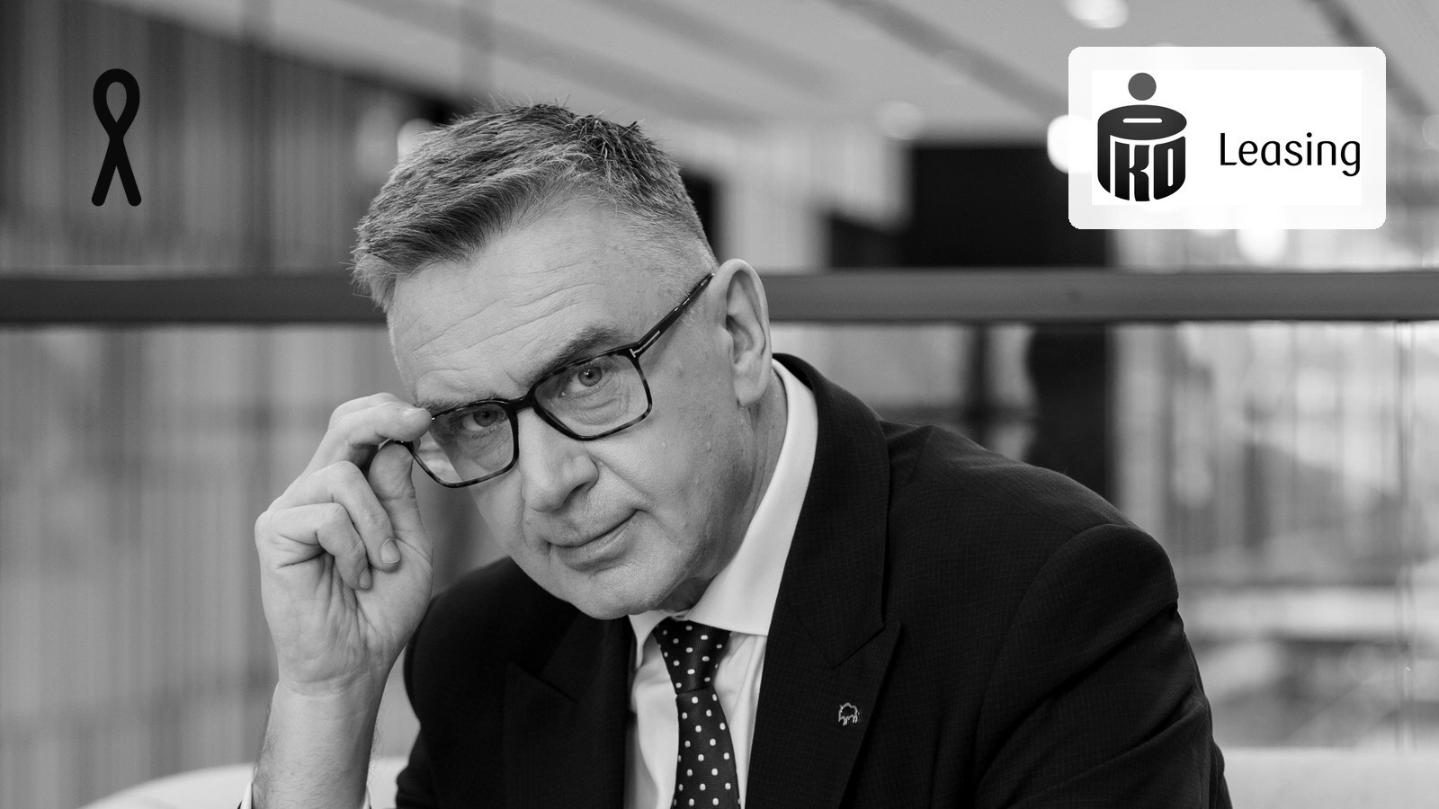 Andrzej Krzemiński w ciemnym garniturze poprawia okulary, siedząc w przeszklonym wnętrzu biurowym