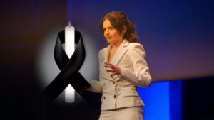 Katarzyna Zaczek w białym garniturze przemawia na scenie, obok zapalona świeca z czarną wstążką