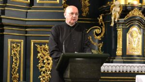 Ksiądz Marek Pieńkowski w czarnej sutannie przemawia przy mównicy przed złoconym ołtarzem kościoła w Jarosławiu