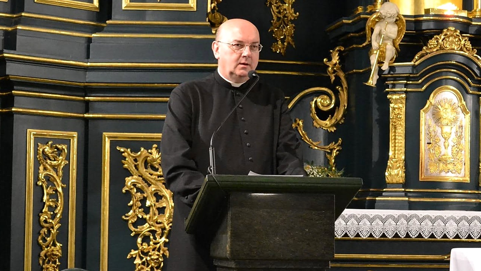 Ksiądz Marek Pieńkowski w czarnej sutannie przemawia przy mównicy przed złoconym ołtarzem kościoła w Jarosławiu