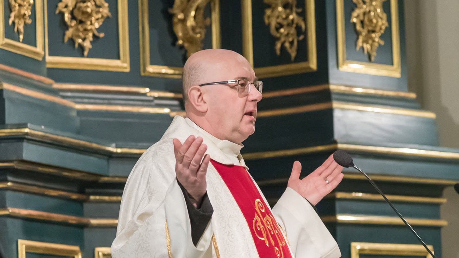 Ksiądz Marek Pieńkowski w białym ornacie z czerwoną stułą głosi kazanie przed złoconym ołtarzem