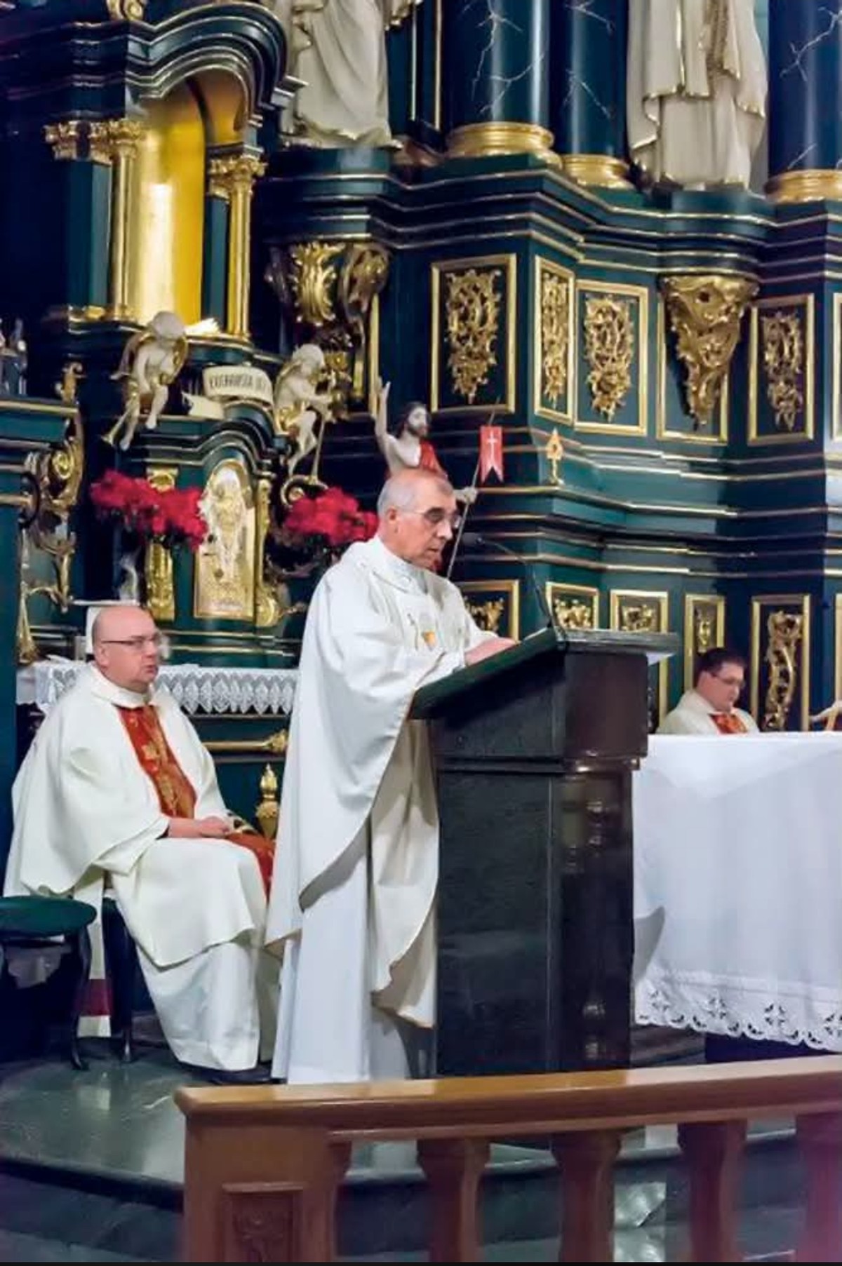 Ksiądz Stanisław Zbojnowicz w białej albie czyta przy ambonie na tle zielono-złotego barokowego ołtarza, obok dwaj koncelebransi