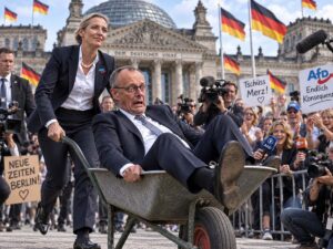 Ilustracja satyryczna: Alice Weidel wiezie kanclerza Friedricha Merza na taczce przed Reichstagiem, w tle transparenty Tschuss Merz