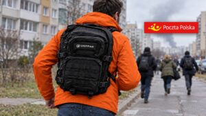Mężczyzna w pomarańczowej kurtce z czarnym plecakiem ewakuacyjnym Defpak oddala się szybko ulicą osiedla, obok logo Poczty Polskiej