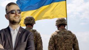 Wicepremier i minister obrony narodowej w ciemnym garniturze, w tle ukraińscy żołnierze przed flagą Ukrainy