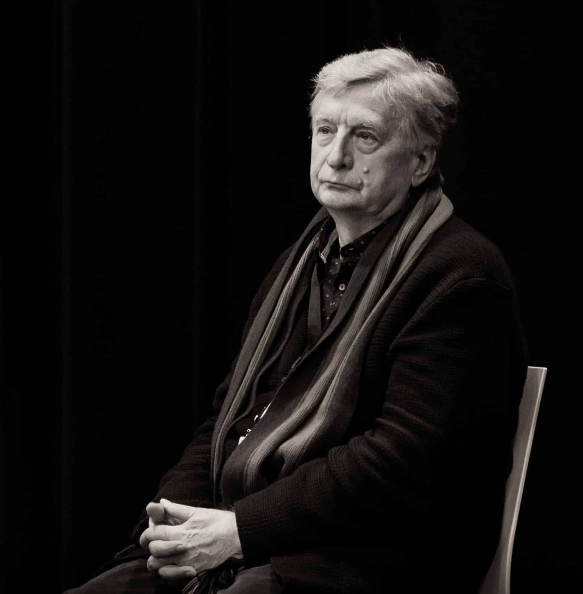 Ryszard Lenczewski w swetrze i szaliku siedzi na krześle na czarnym tle, portret studyjny
