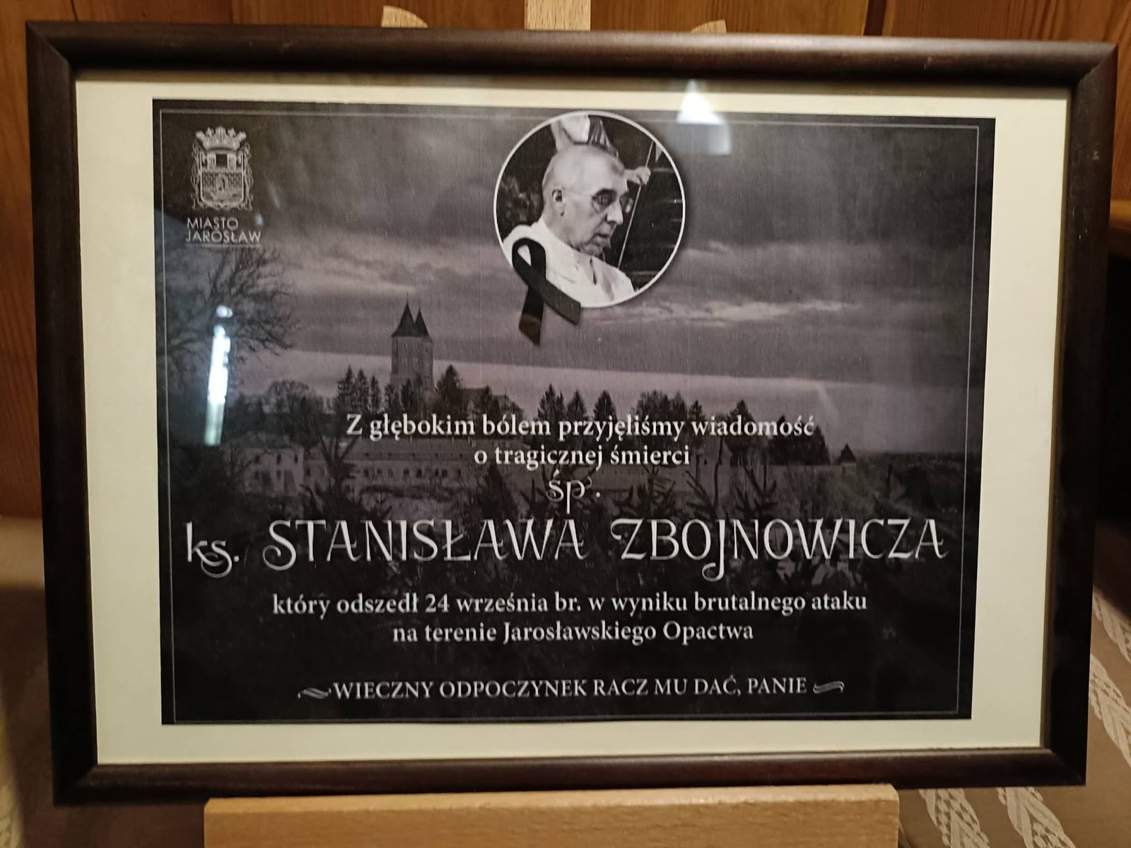 Oprawiona tablica Miasta Jarosław z portretem ks. Stanisława Zbojnowicza i czarną wstążką na tle opactwa