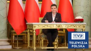 Marta Cienkowska podpisuje dokument na tle biało-czerwonych flag, w rogu kadru znak Telewizji Polskiej