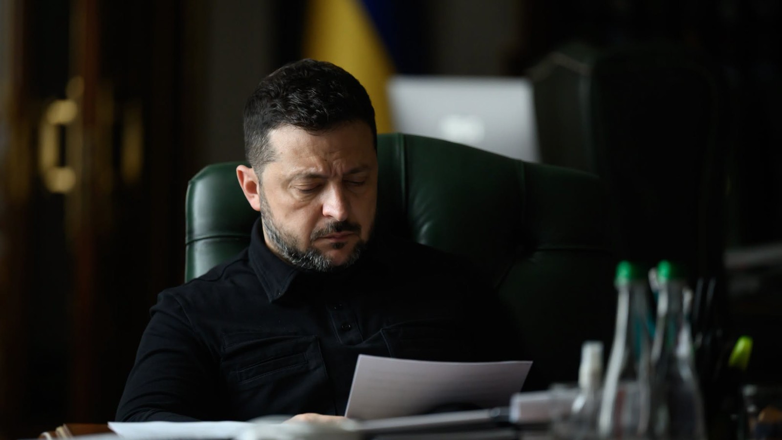 Wołodymyr Zełenski czyta dokumenty przy biurku w gabinecie z ukraińską flagą w tle