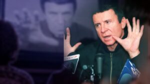 Anatolij Kaszpirowski z uniesionymi dłońmi przy mikrofonach, w tle telewizor z jego seansem i widzowie