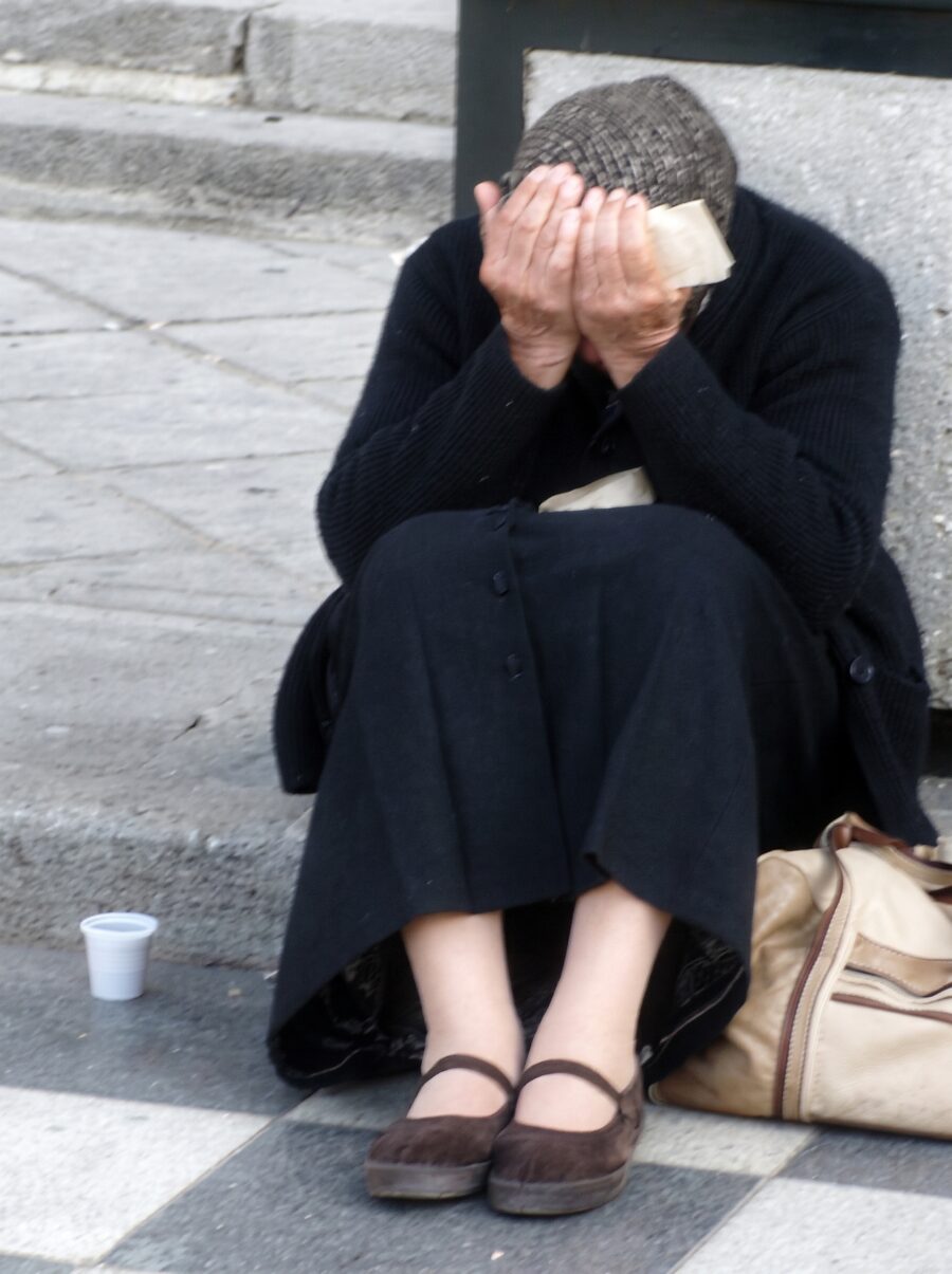 Bezdomna kobieta w ciemnym płaszczu siedzi na chodniku z twarzą ukrytą w dłoniach, obok kubek i torba