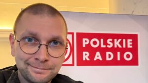 Ks. Mateusz Szerszeń CSMA w koloratce na tle ścianki Polskiego Radia, uśmiechnięty, w okrągłych okularach