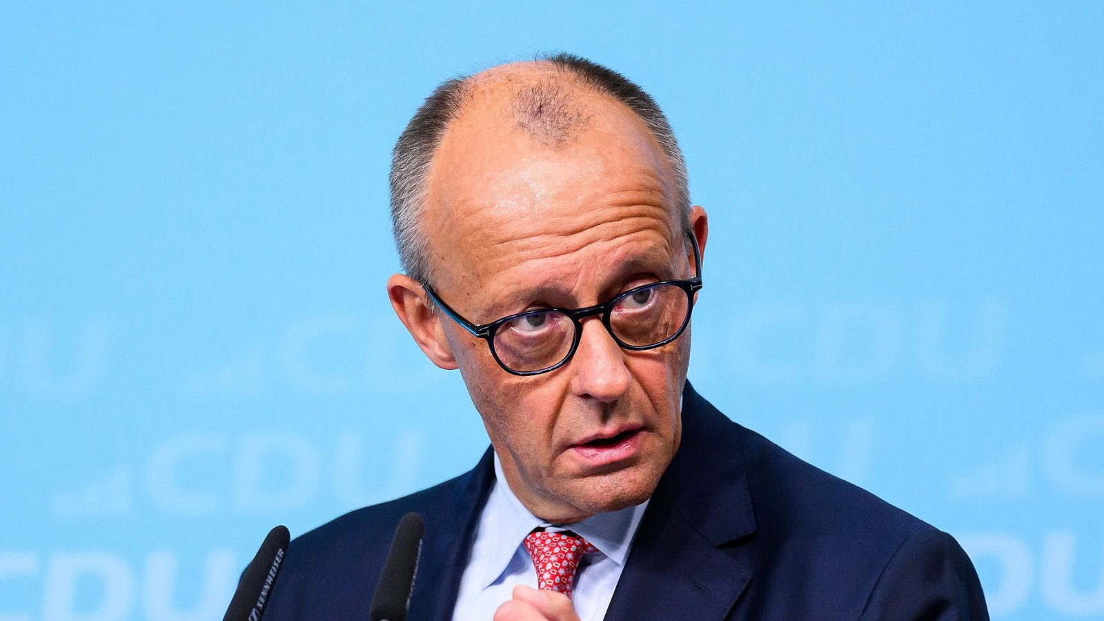 Friedrich Merz w okularach i czerwonym krawacie przemawia przy mównicy na tle niebieskiej ścianki z logo CDU