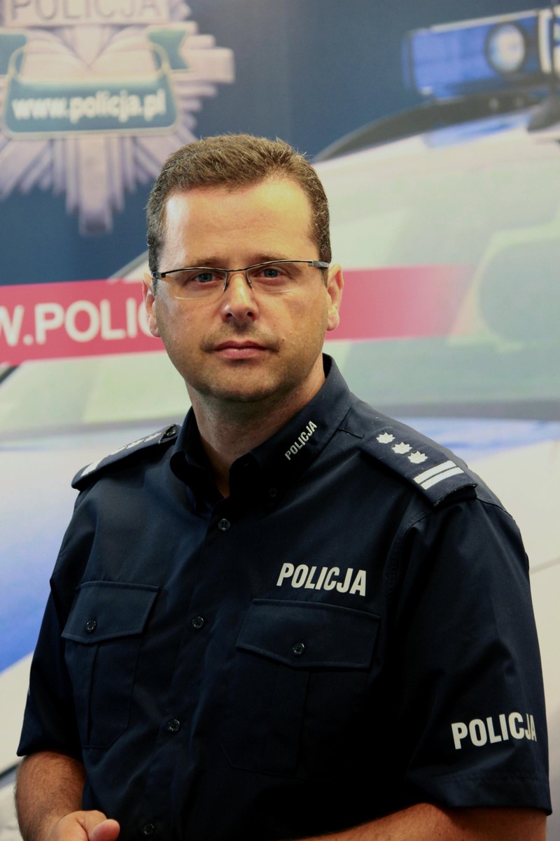 Mariusz Sokołowski w mundurze policyjnym na tle ścianki z logo Policji i radiowozem
