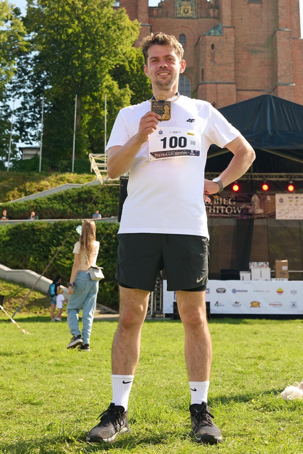 Michał Kołodziejczak w stroju biegowym z numerem 100 pokazuje medal Biegu Lechitów przed katedrą w Gnieźnie