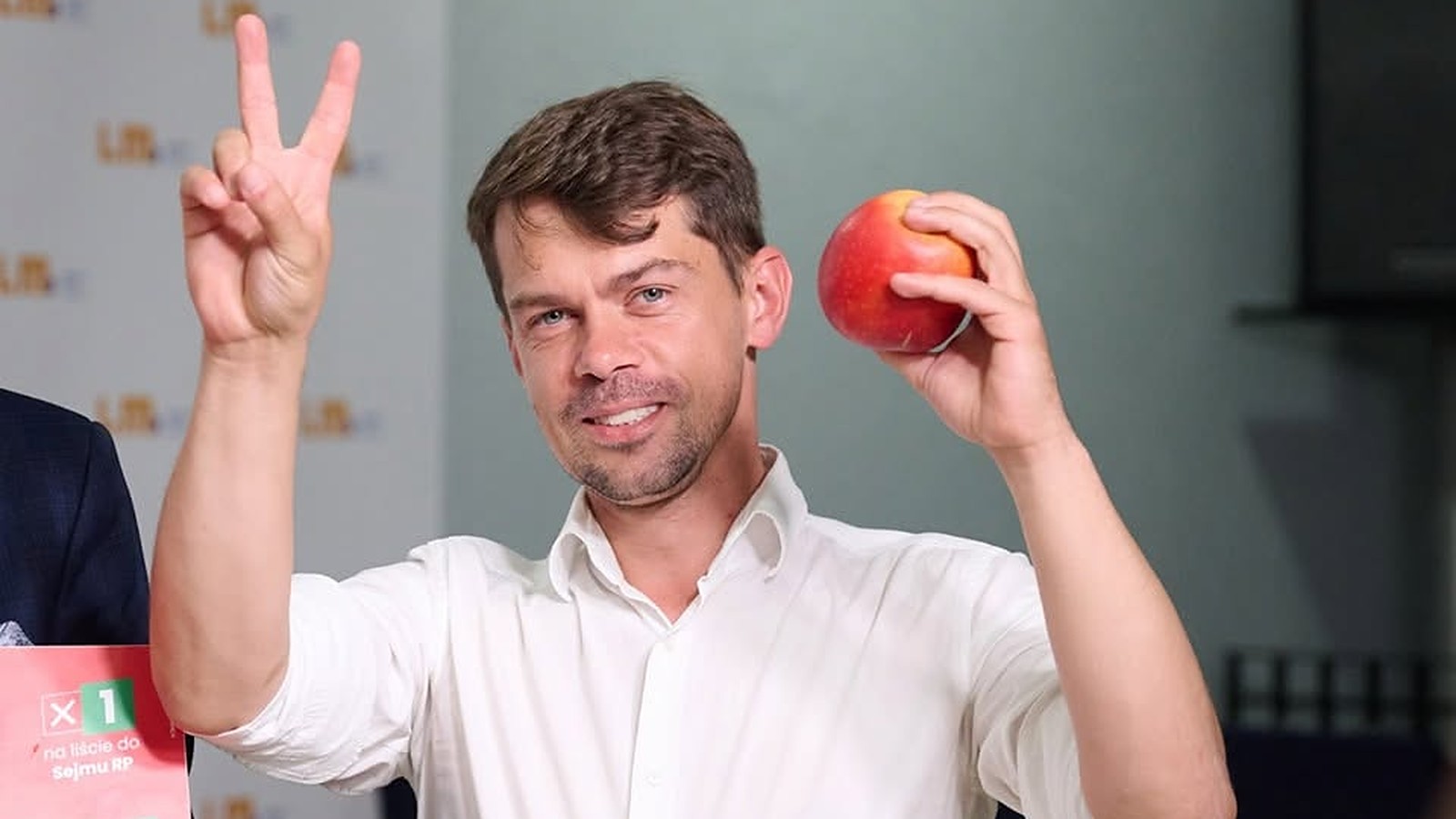 Michał Kołodziejczak, poseł w wieku 38 lat, w białej koszuli pokazuje znak V i trzyma czerwone jabłko