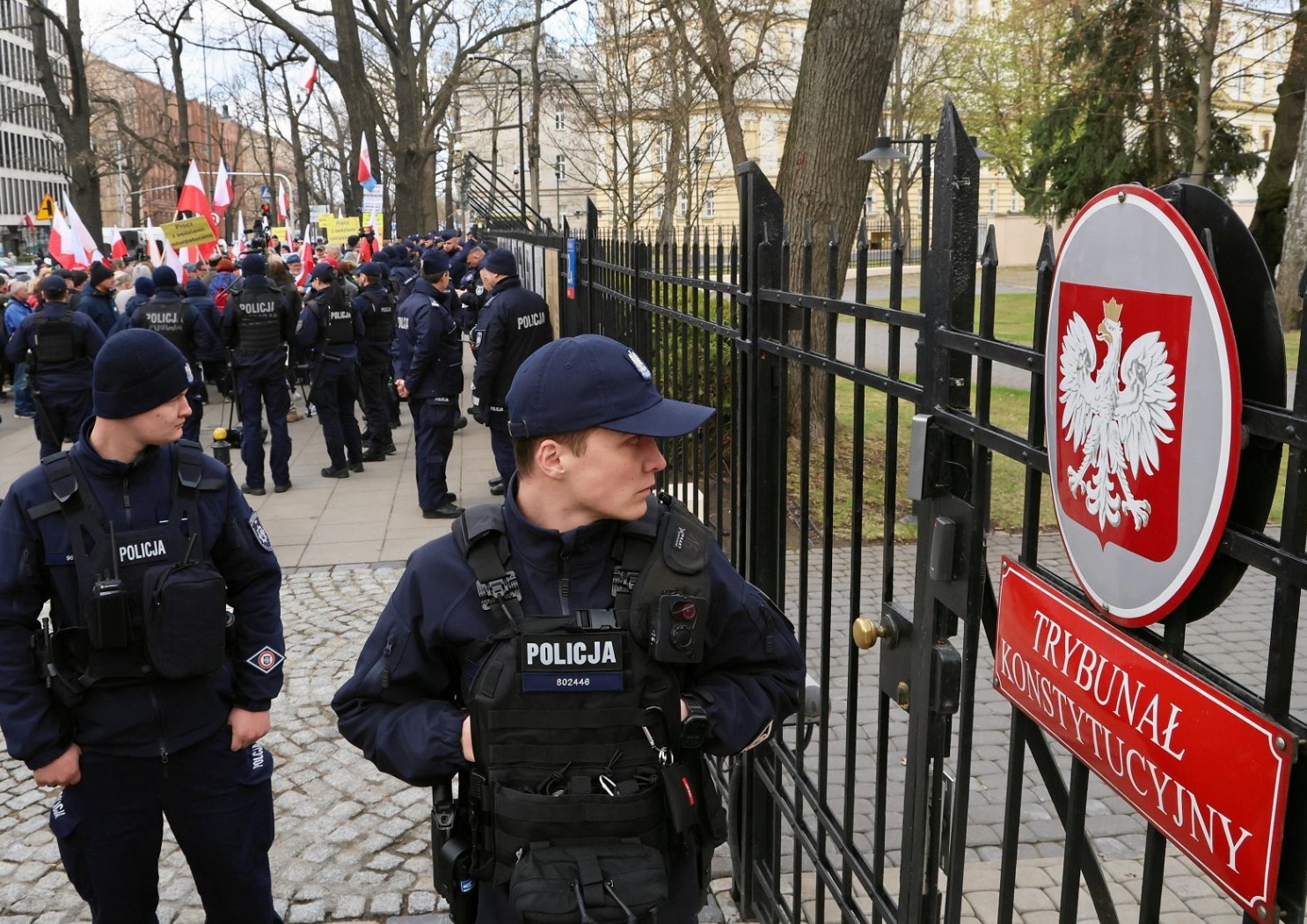 Kordon policjantów w kamizelkach przed bramą Trybunału Konstytucyjnego, w tle demonstranci z flagami