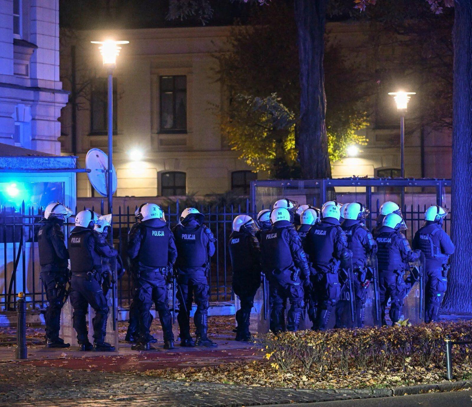 Policjanci w kaskach i kamizelkach z tarczami stoją nocą przy ogrodzeniu siedziby Trybunału Konstytucyjnego