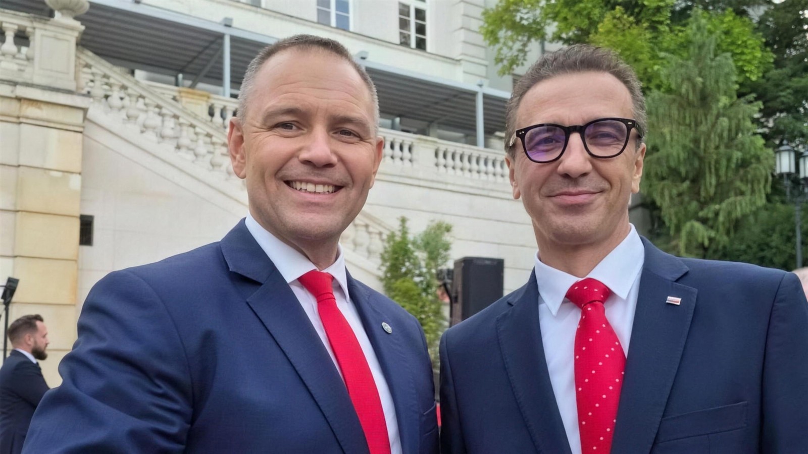 Poseł Jarosław Sachajko w granatowym garniturze i czerwonym krawacie podczas spotkania przed pałacowym budynkiem