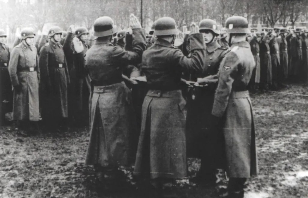 Żołnierze SS Galizien w hełmach i płaszczach składają przysięgę z uniesioną prawą ręką na placu