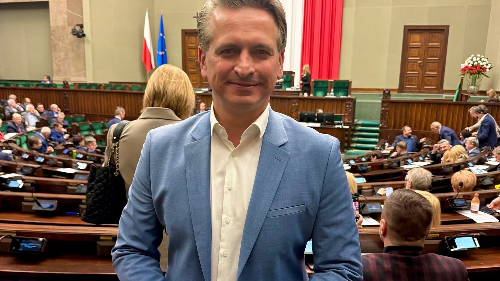 Robert Wardzała w jasnoniebieskiej marynarce na sali plenarnej Sejmu, w tle godło i posłowie w ławach