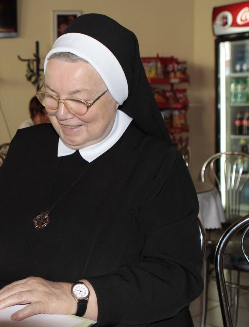 Uśmiechnięta siostra Anastazja Pustelnik w habicie i okularach wpisuje dedykację do książki przy stoliku