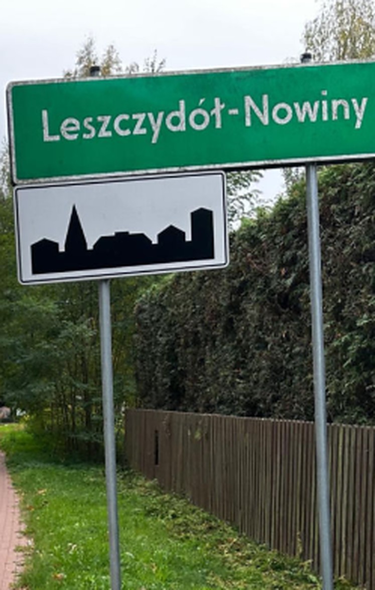 Zielona tablica z nazwą wsi Leszczydół-Nowiny przy drodze, w tle drewniany płot i drzewa