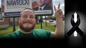 Uśmiechnięty Waldemar Krysiak w zielonej koszulce pokazuje znak zwycięstwa, obok świeca z czarną wstążką
